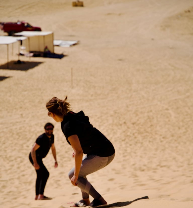 A person sandboarding run down a sand dune. Another person, standing in the background.