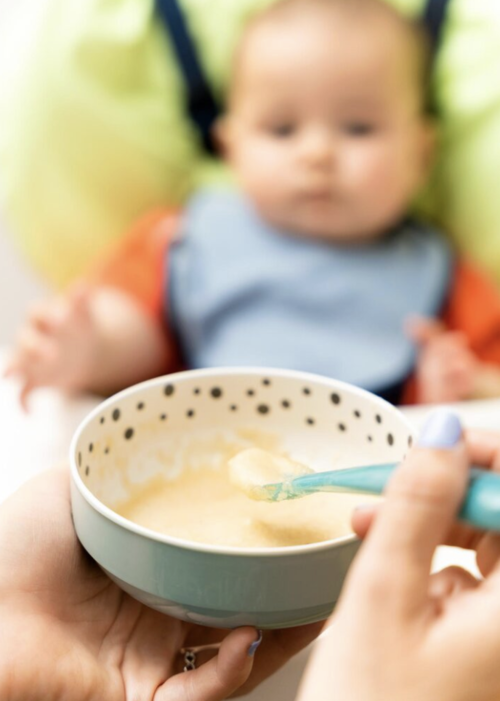 madre alimenta a un bebé con puré orgánico en una silla alta usando una cuchara azul.