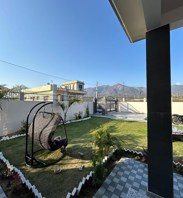 A modern outdoor patio and green garden featuring a wicker hanging egg chair and mountain views.