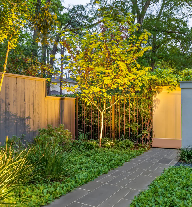 Backyard landscape lighting illuminating a stone path, lush garden beds, and a privacy fence at dusk.