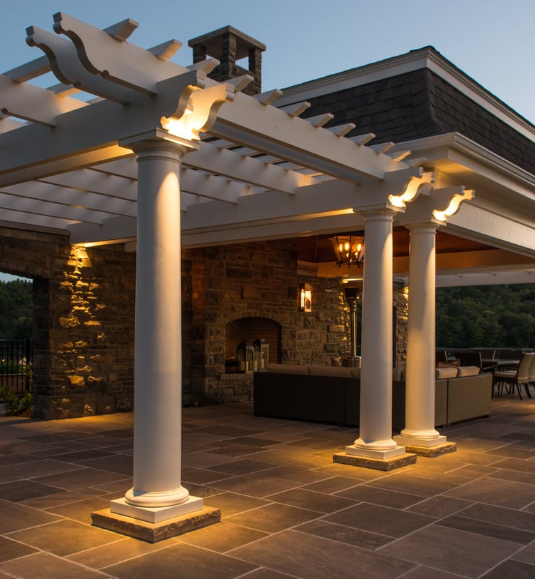 Luxury outdoor patio with stone fireplace, lit white pergola, and bluestone pavers at dusk.
