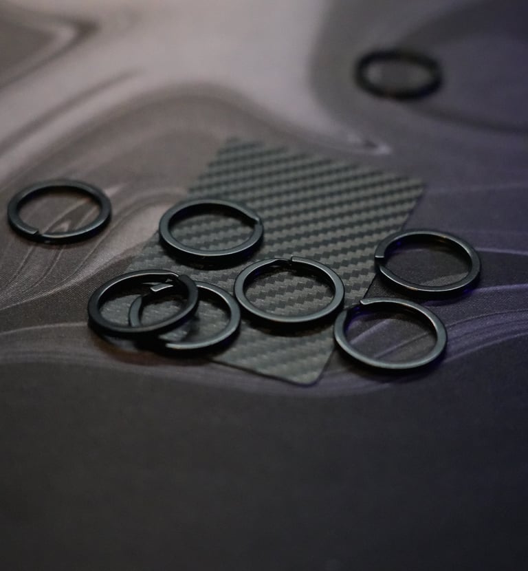 Small black circular split rings and metal fasteners for EDC gear on a carbon fiber mat.