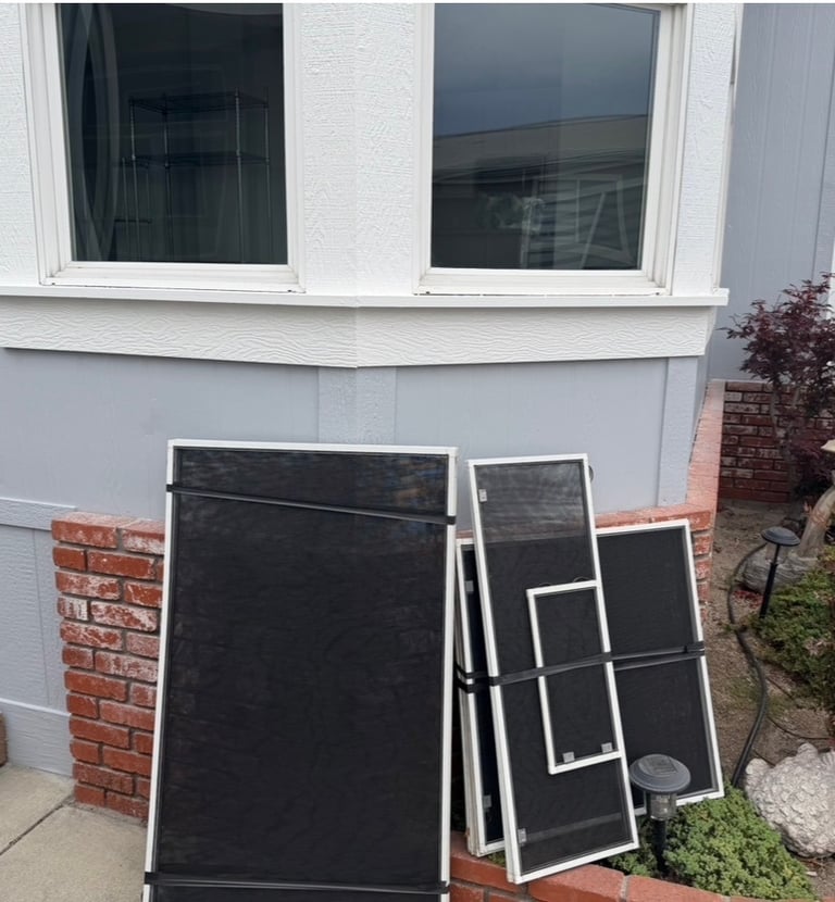 Replacement window screens leaning against a mobile home exterior with brick accents.