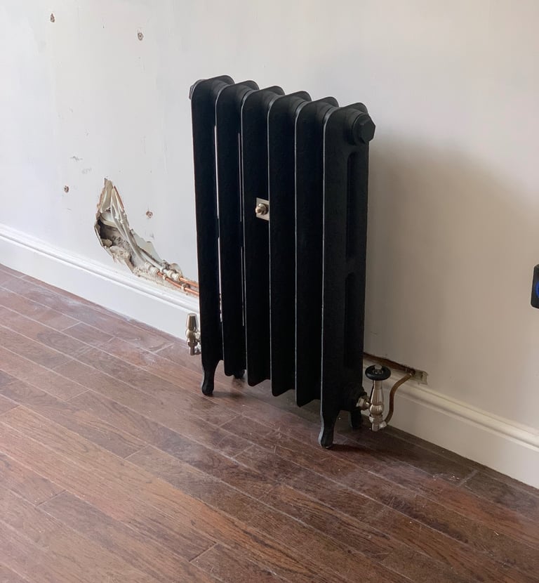Black traditional cast iron radiator installed against a white wall with exposed copper piping.