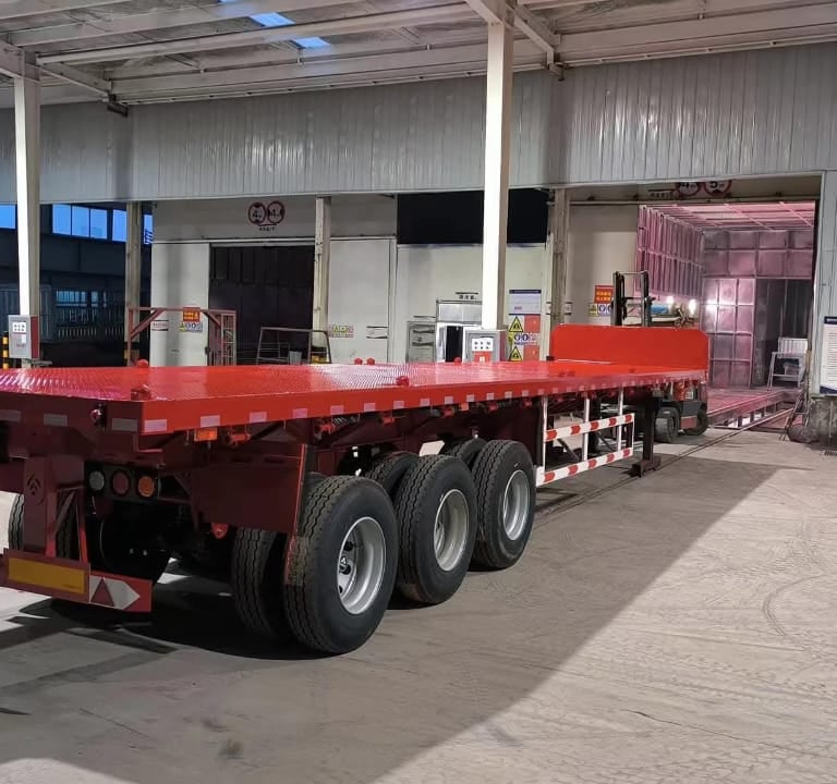 Red 3-axle flatbed semi-trailer parked in our industrial manufacturing warehouse facility.