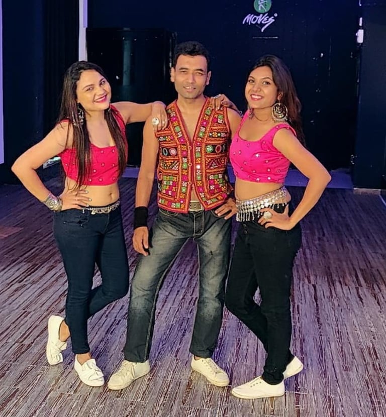 Three Bollywood dancers pose in a studio wearing colorful traditional embroidered vests and pink crop tops.