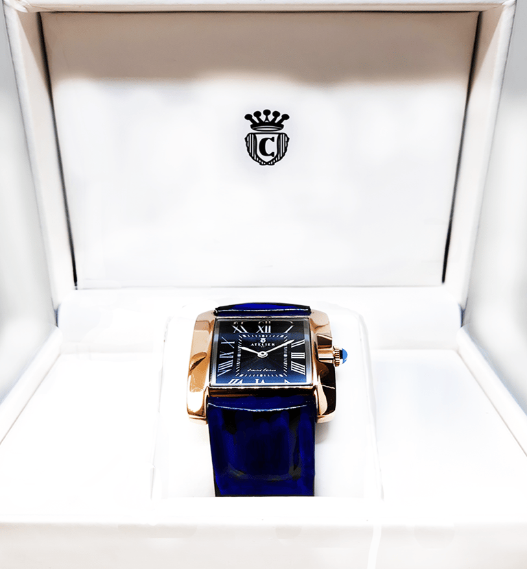 Luxury rectangular rose gold watch with a blue leather strap and Roman numeral dial in a white display box.