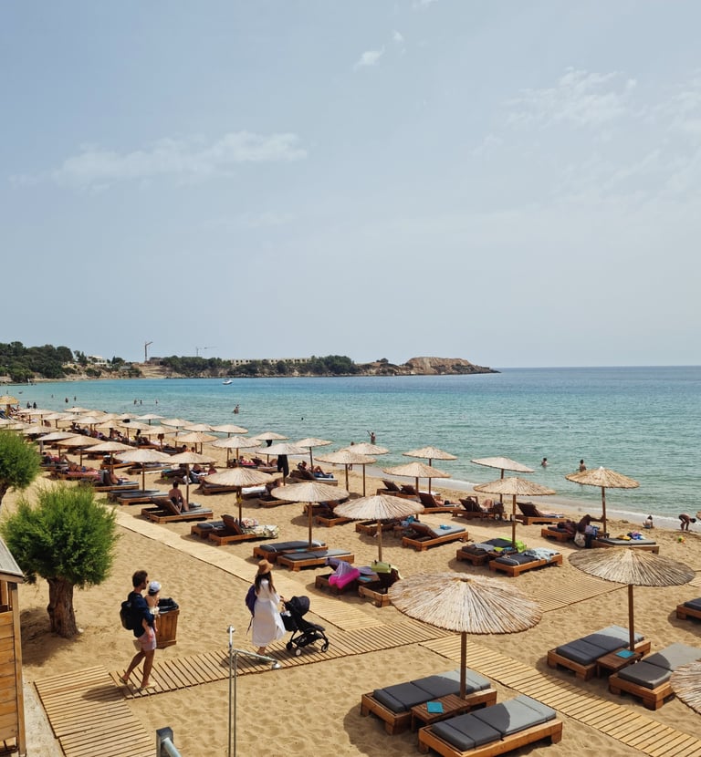 Luxury beach club with rows of thatched straw umbrellas and wooden sun loungers on a sandy Greek shore.