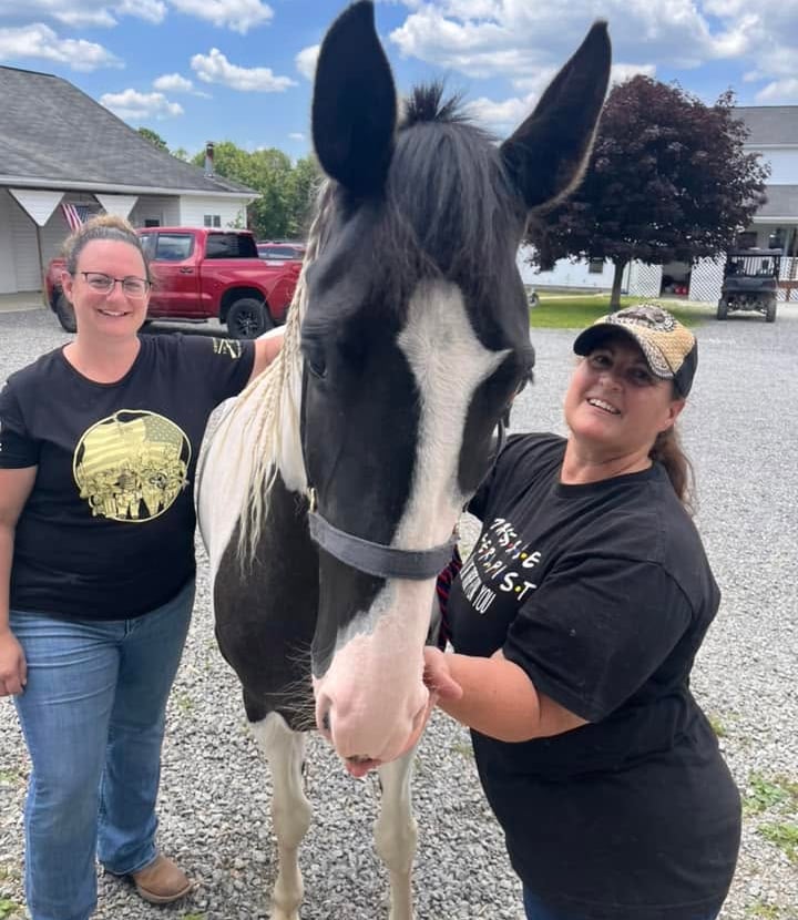 The owners standing outdoors next to a black and white spotted horse with a braided mane.