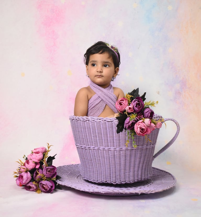 Baby girl posing in a purple wicker teacup basket with floral decorations for a studio photoshoot.
