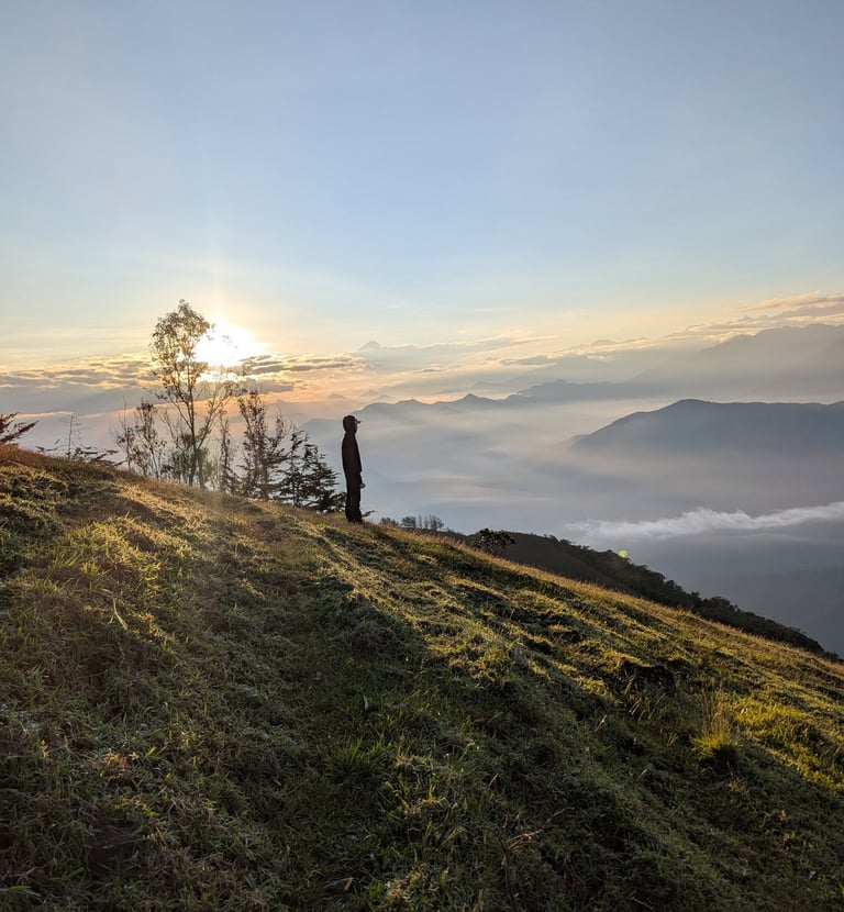 A hiker stands on a grassy mountain ridge watching the sunrise over a foggy valley.