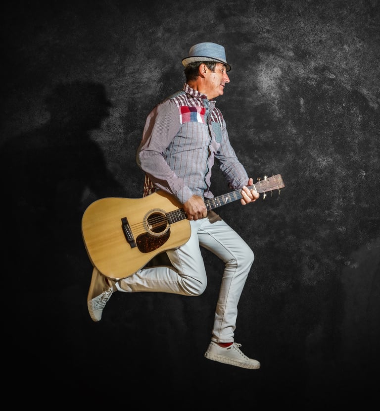 Joël Royer en photo prise de profil en train de sauter avec un chapeau sur la tête et tenant une guitare sèche dans ses mains