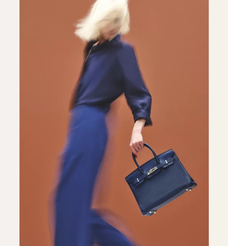 Model in motion holding an Hermes Birkin navy blue luxury handbag against a vibrant backdrop.