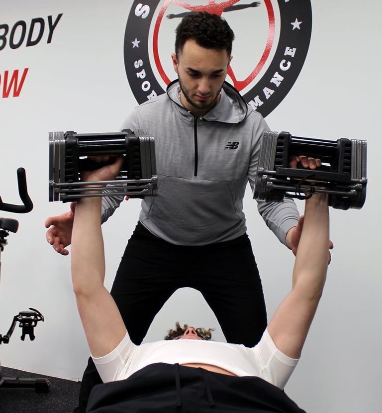 A personal trainer spots a man performing a chest press with adjustable dumbbells in a gym.