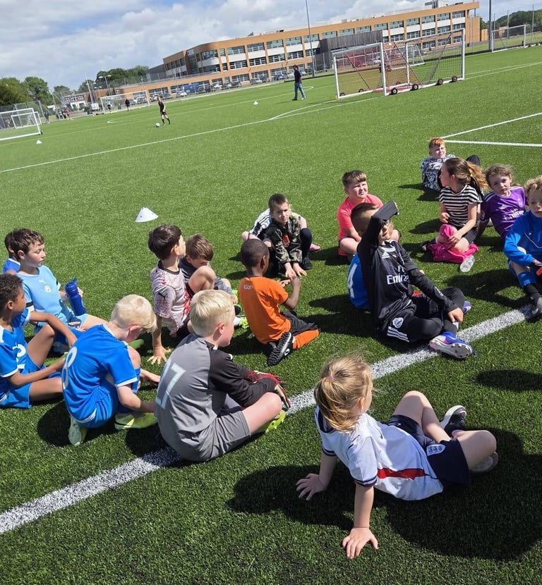 A group of young children sitting in a circle on a green artificial turf football pitch