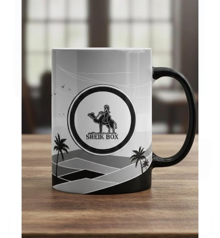 White ceramic coffee mug with black handle featuring a desert landscape and Sheik Box camel logo.