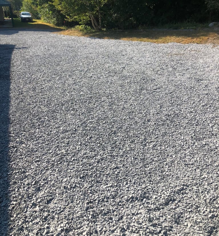 A wide gray crushed stone gravel driveway leading past green trees to a parked car.
