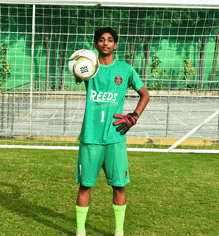 A youth football goalkeeper in a green kit holding a soccer ball in front of a goal post on a grass pitch.