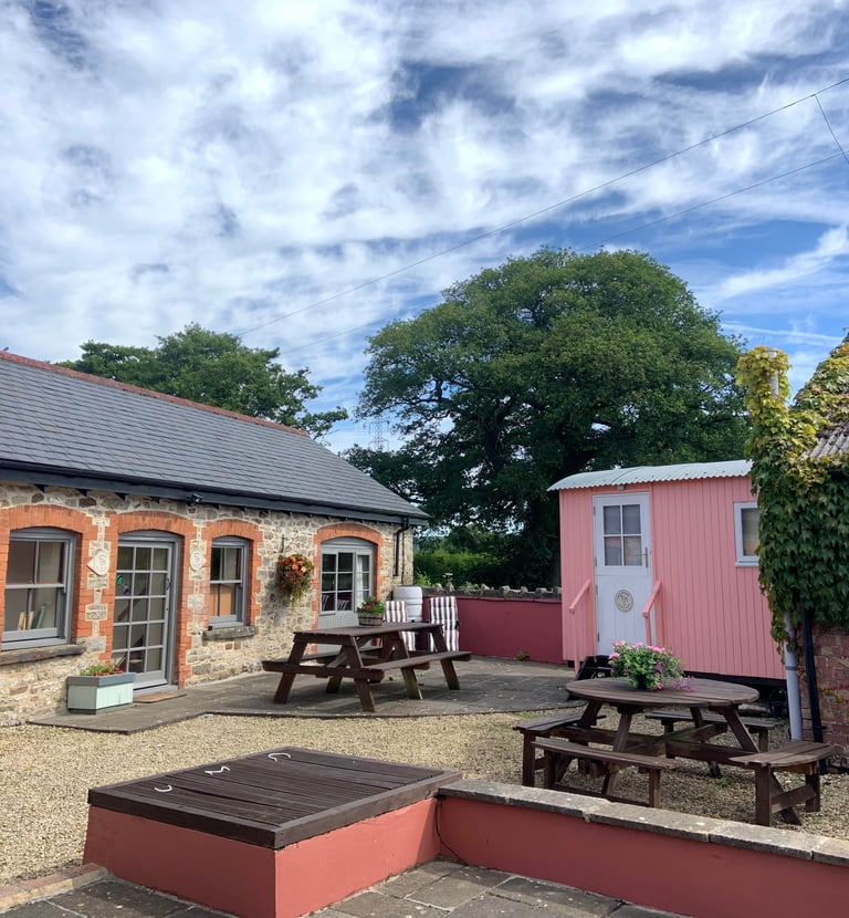 Rustic stone cottage and pink shepherd hut with outdoor wooden picnic tables on a sunny gravel patio.