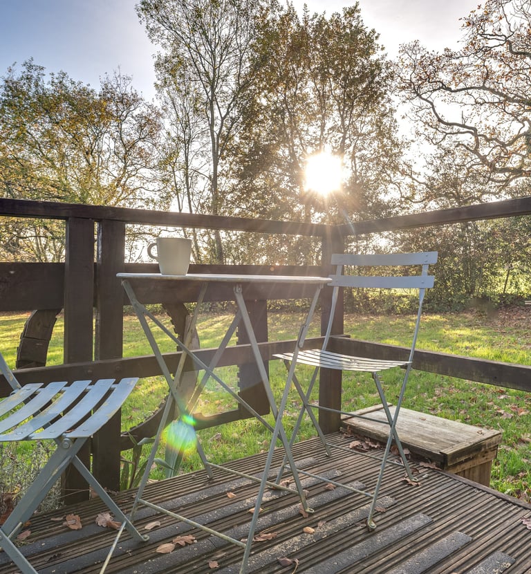 Metal bistro set on a wooden garden deck with golden hour sunlight filtering through autumn trees.