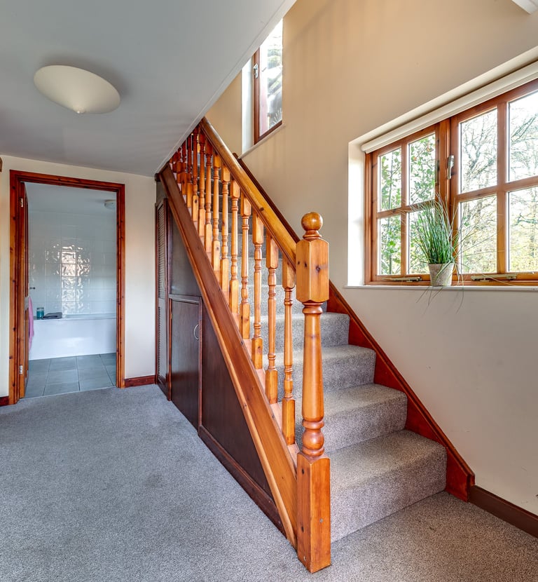 Home hallway featuring grey carpet, wooden staircase with spindles, and a view into a white bathroom.