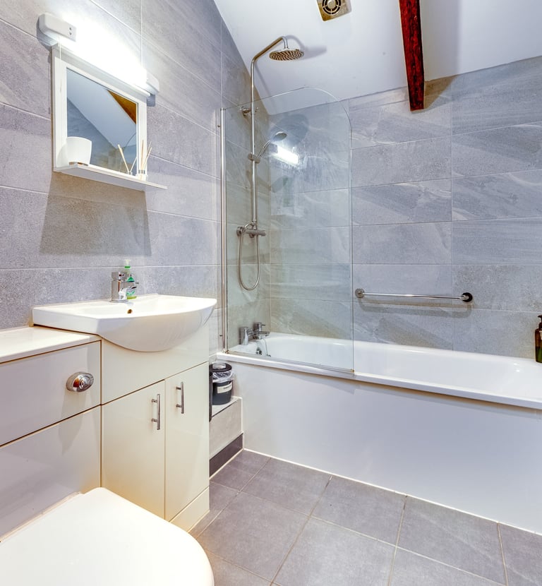 Modern bathroom with grey stone tiles, a white bathtub with a glass shower screen, and a vanity sink.
