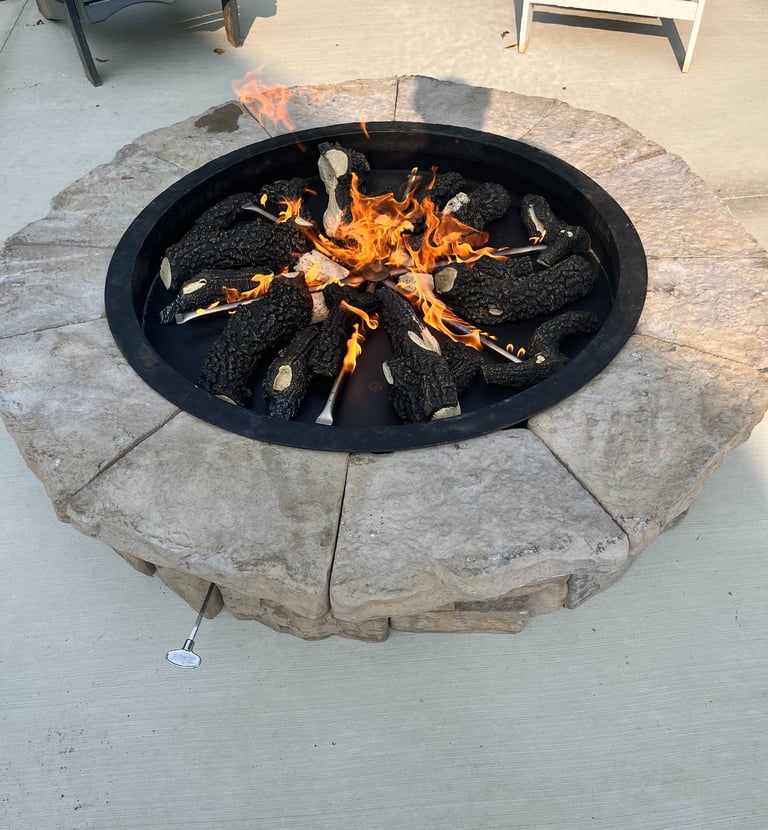 A round outdoor stone gas fire pit with burning ceramic logs on a concrete patio.