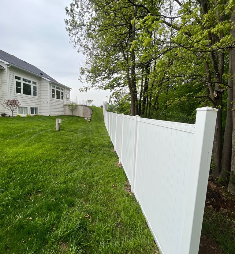 White vinyl privacy fence installed in a grassy backyard next to a residential home and green trees.