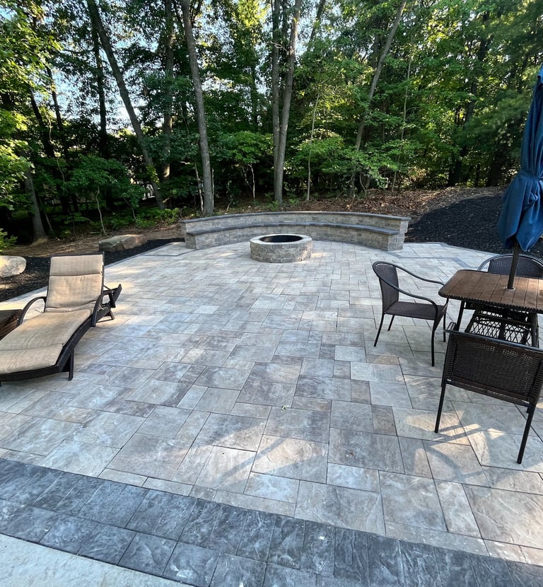 Luxury backyard stone paver patio featuring a round fire pit, built-in stone bench, and outdoor lounge furniture.