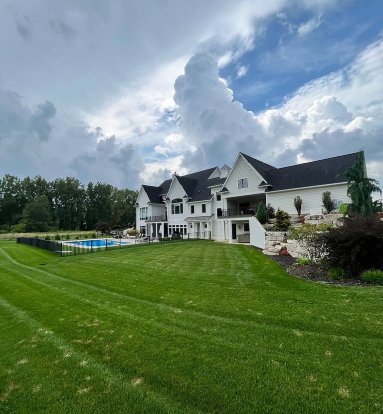 Modern white farmhouse mansion with a luxury backyard pool and manicured green lawn under a cloudy sky.