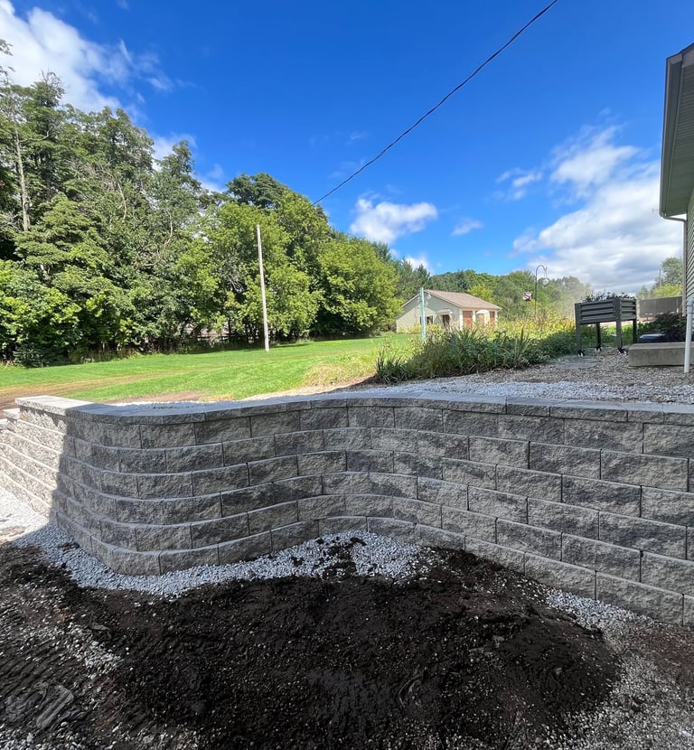Newly constructed curved gray stone retaining wall in a backyard landscape with green lawn and trees.