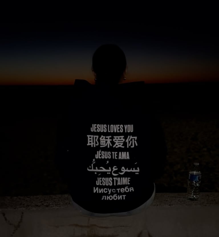Silhouetted person at sunset wearing hoodie reads Jesus loves you in many languages worldwide faith!