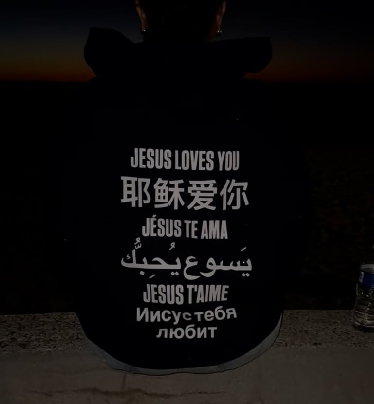 Silhouetted person at sunset wearing a hoodie that reads Jesus loves you in many languages.
