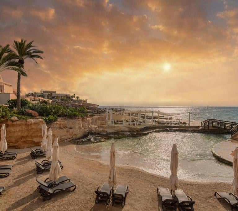 Luxury beach resort with sun loungers and palm trees at sunset overlooking a scenic ocean cove.