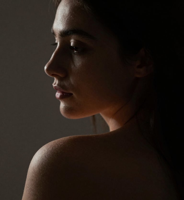 A fine art portrait in extreme chiaroscuro. The light softly catches the curve of a shoulder and the profile of a face, leaving the rest in deep #1F1E1C shadows. Warm, intimate atmosphere with refined textures.