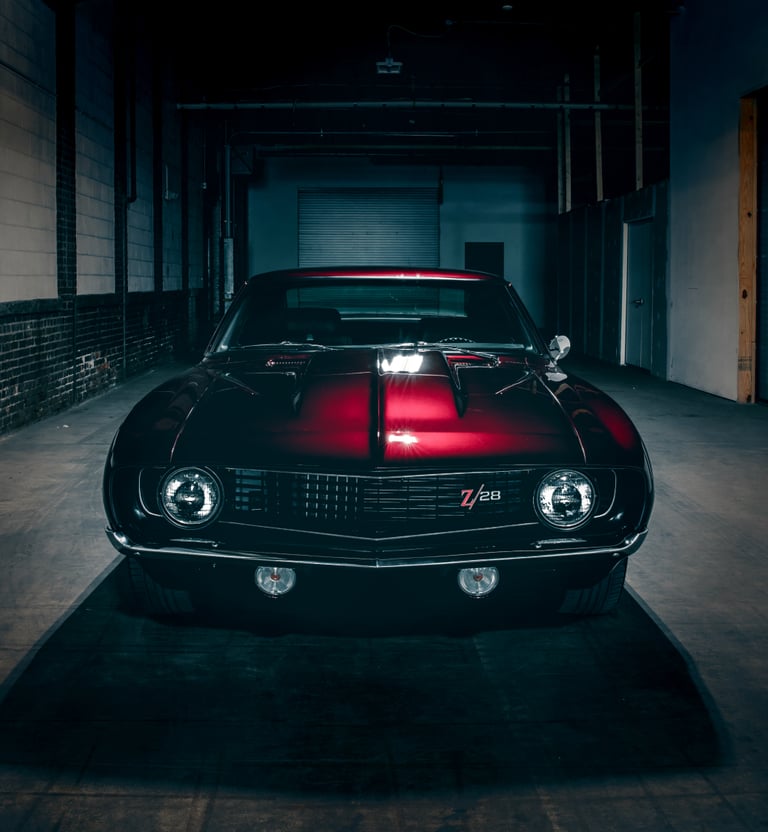 Front view of a classic red and black Chevrolet Camaro Z28 parked in a dark industrial garage.