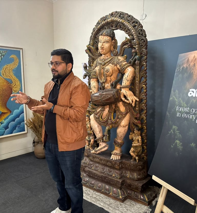 A man presenting next to an ancient Indian wooden sculpture and Atavi forest brand signage in an art gallery.