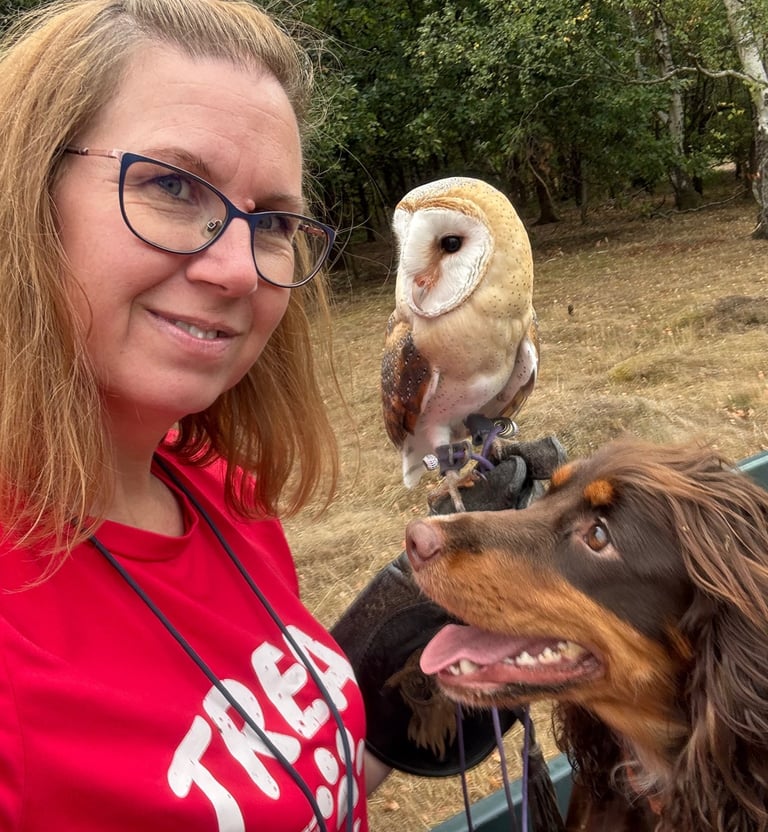 Dog trainer smiling with her spaniel and a barn owl perched on her fist outdoors.