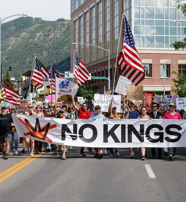 Protesters march through a city street holding a large No Kings banner and American flags during a political rally.