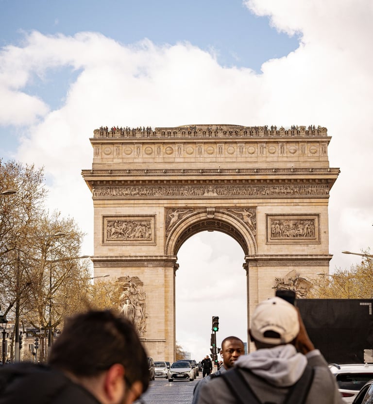 Tourists taking photos of the Arc de Triomphe monument in Paris under a blue sky.