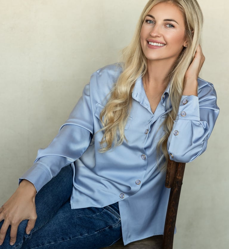 Moteris koučerė sėdi ant rudos kėdės, šypsosi, pasirėmusi ranka galvą ir dėvinti žydrus marškinius