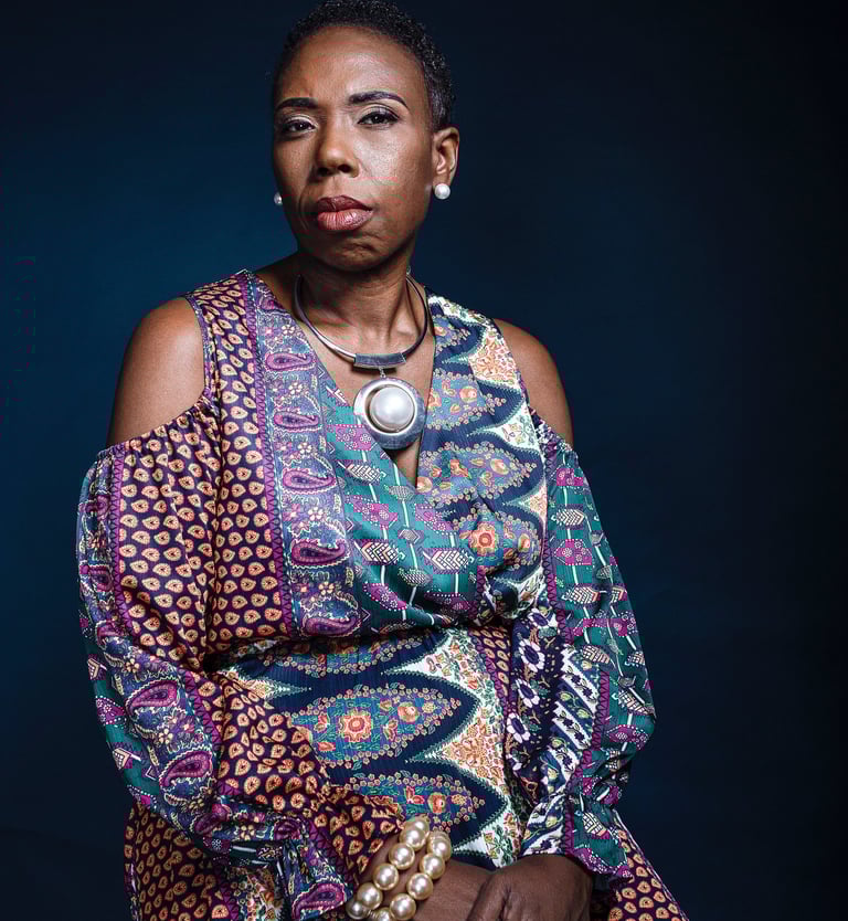 Portrait of a Black woman in a colorful patterned dress and pearl jewelry against a dark background.
