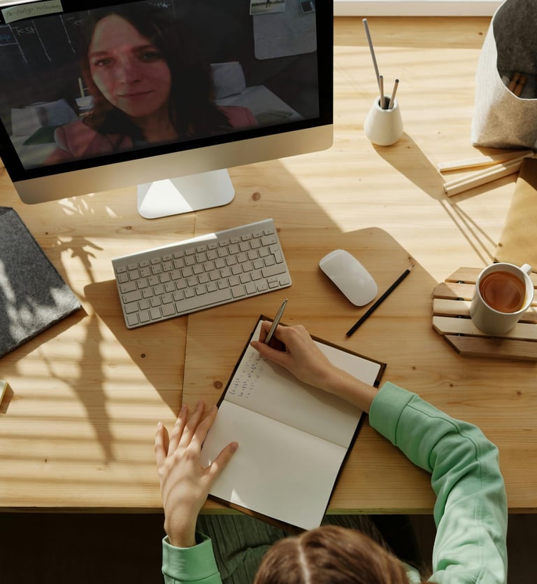 A woman takes notes in a notebook during a remote video conference call on her desktop computer.