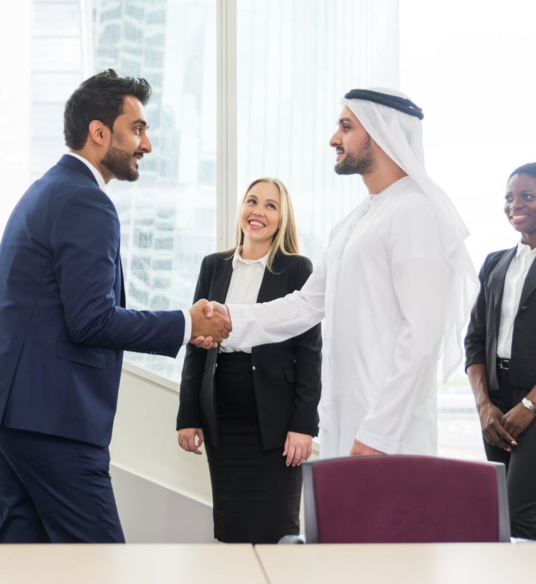 Businessman in a suit shakes hands with another in Emirati attire, while a businesswoman smiles