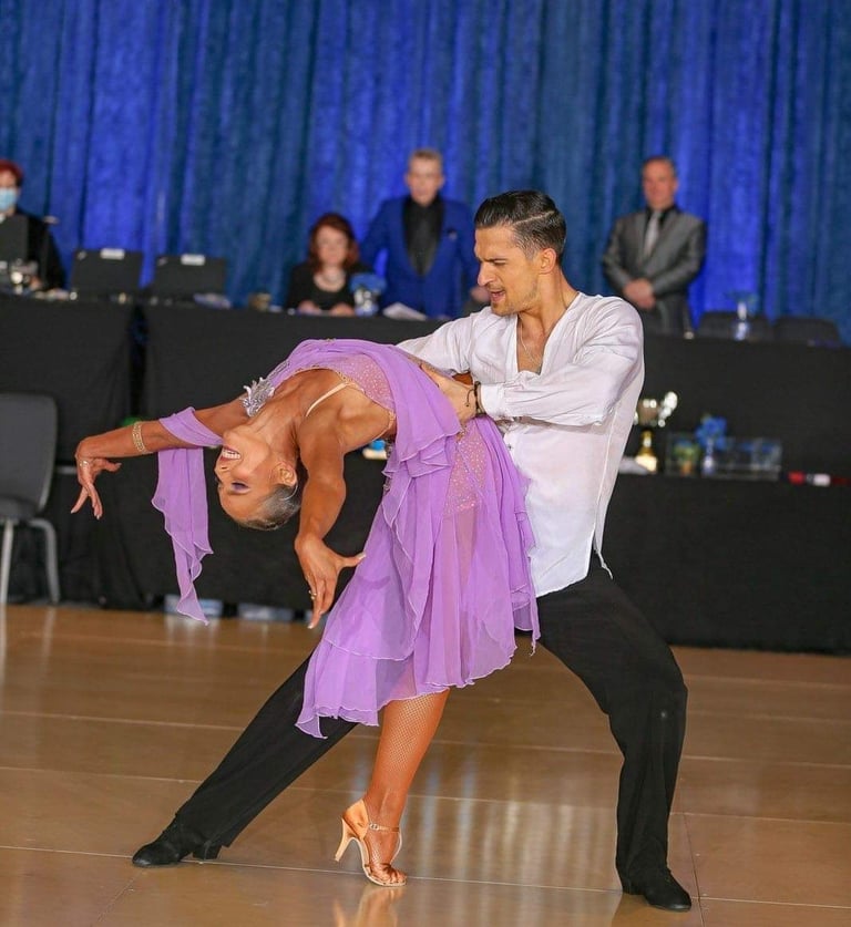 A man in a white silk shirt holds a woman as she arches her back, wearing a purple silk dress