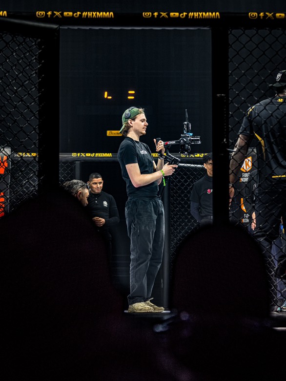 Professional videographer filming an MMA match with a gimbal stabilizer inside a dark octagon cage.