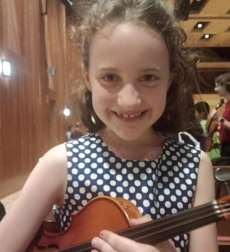 A smiling young girl in a polka dot dress holds a violin during a summer Suzuki Institute.