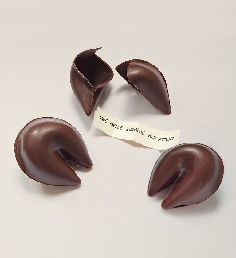 Handmade dark chocolate fortune cookies on a white surface with a paper message in French.