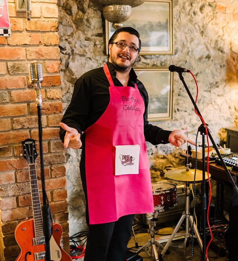 a man dressed in 1950s costume for a themed murder mystery. He is smiling and wearing a pink apron.
