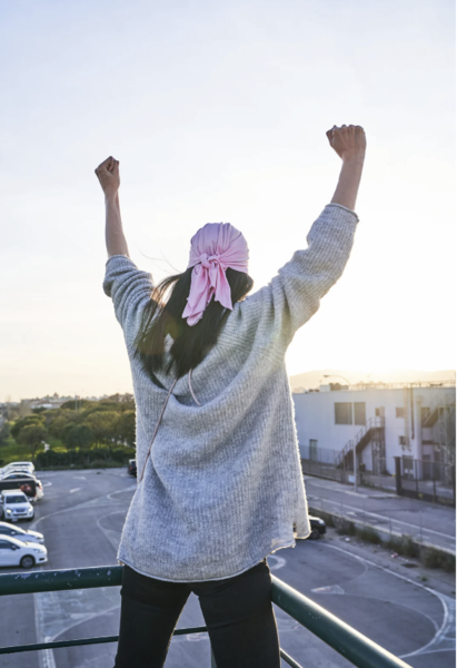 A breast cancer survivor in a pink headscarf raising fists in victory on a sunny balcony.